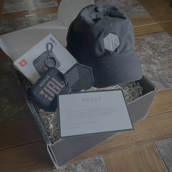 Photograph of a gift box: on top of crinkled, biodegradable packing materials is a GO4 JBL speaker marked with the word "NEXUS". There is a black ballcap with the hexagonal Nexus logo on its front, a sticker of the Nexus logo, and a thank-you card from Nexus for participating in Nexus' testnet.