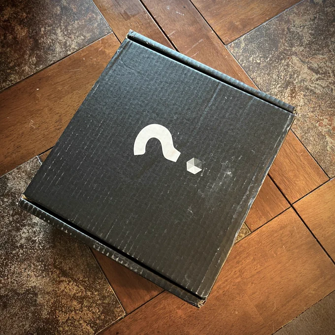 Photo of a cardboard shipping box. It is weathered black cardboard, with a question mark printed in white. The dot of the question mark is the Nexus hexagonal logo.
