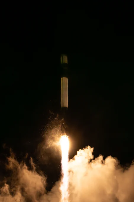 The photo captures the PREFIRE launch at night. A bright, fiery plume of exhaust illuminates the scene, casting light on billowing clouds of smoke and steam. The Electron rocket's silhouette is visible against the dark sky, with a faint glow highlighting its structure as it ascends. The contrast between the intense light of the engines and the surrounding darkness creates a dramatic effect.