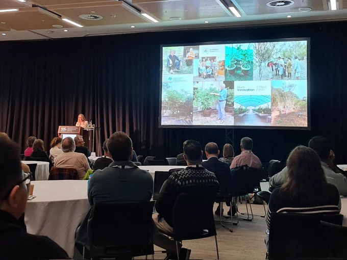 Dr Nerida Donovan presenting a slide of people involved in past citrus work in front people seated around round tables. 