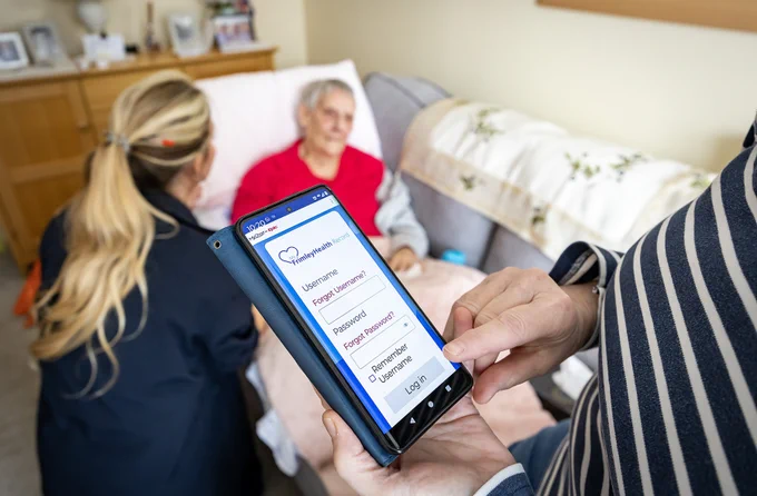 An individual displays a healthcare app on a smartphone screen to another person seated on a sofa, while an elderly person sits in the background.