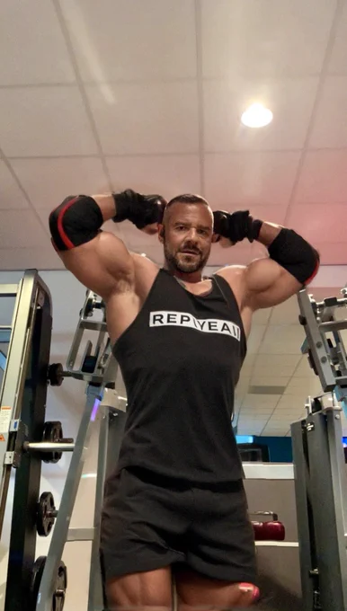 A muscular man in a gym posing with both arms flexed behind his head, wearing a tank top that says ‘REPWEAR’, looking confident and ready to scare the protein out of the weights.