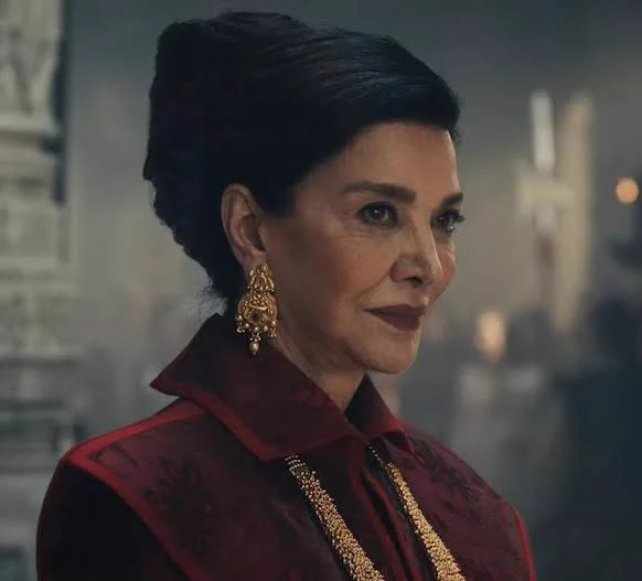 A screenshot of Shohreh Aghdashloo as Elaida in The Wheel of Time.  She is wearing a red dress and gaudy, golden jewelry.  Her expression is superior and calculating as she looks at Siuan on the Amyrlin Seat. 