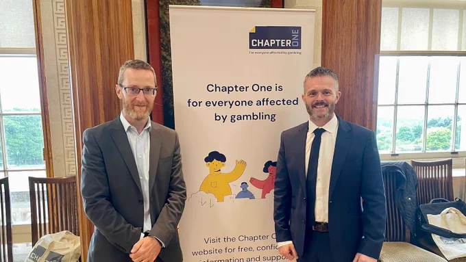 Philip McGuigan MLA and Robbie Butler MLA stand beside a banner with the Chapter One logo. The banner says "Chapter One is for everyone affected by gambling" and "Visit the Chapter One website for free, confidential information and support". 