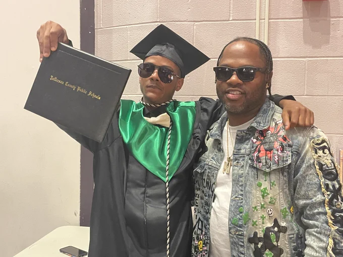 A graduate poses for a picture with another individual and holds up his diploma.