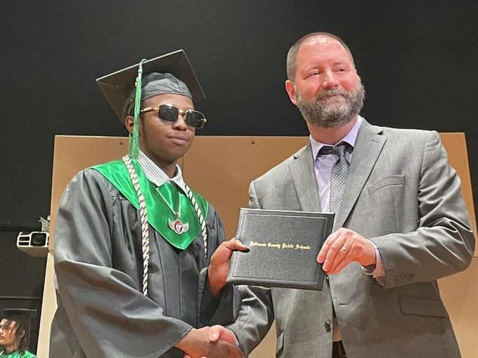 A graduate poses for a picture with a school administrator. Both hold a diploma. 