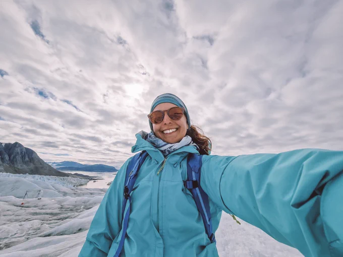 A woman takes a selfie on a frozen landscape wearing a teal jacket, blue book bag, teal headband, and tan sunglasses. The sky above her is covered in white, fluffy clouds. The white ground and sky are the a perfect contrast to her bright blue outfit. 