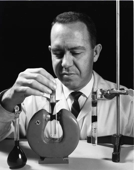 A black-and-white image of NASA researcher Stephen Papell, the inventor of ferrofluids. He wears a lab coat and works with various vials and scientific equipment on a table. Credit: NASA/John Marton