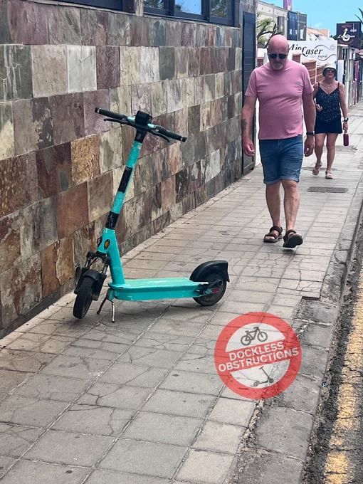 An abandoned e-scooter obstructing a narrow pavement opposite a docking station on the Canarian Island of Fuerteventura.
