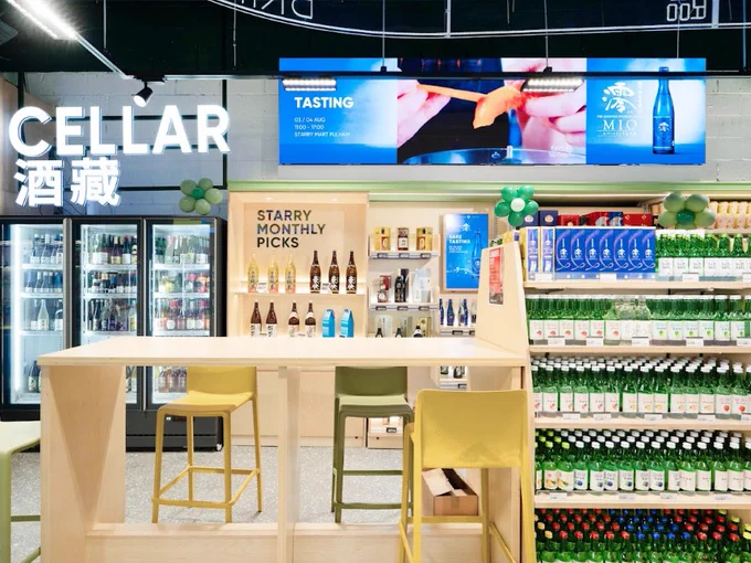 The Cellar counter at a Starry Mart store, featuring a variety of drinks displayed on shelves, a tasting area with a digital screen, and a seated bar area with stools.
