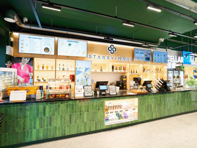 Interior view of Starry Mart, featuring a food counter with various menu displays, serving equipment, and a cheerful decor with distinctive green tile. A digital screen shows promotions and a staff member appears behind the counter.