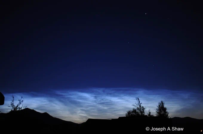A photo showing glowing noctilucent clouds over a dark sky, with a mountain silhouette and rooftops in the foreground. The clouds have a blue-silvery hue, typical of noctilucent clouds. Photo taken from Bozeman, MT on 16 July 2009. Copyright: Joseph A Shaw. 