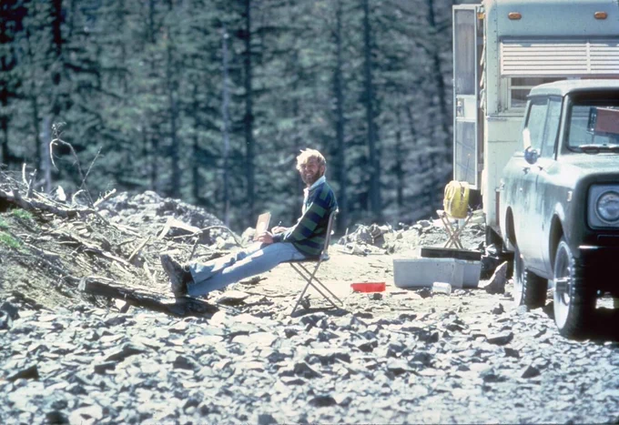 David Johnston in blue and green striped shirt sitting on camping char on road cobble near camper and truck with trees in background. 
