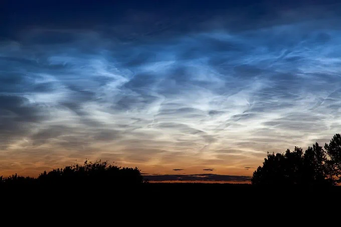 A sunset sky with wispy, glowing clouds in shades of blue and white above a dark silhouette of trees and bushes on the horizon.

