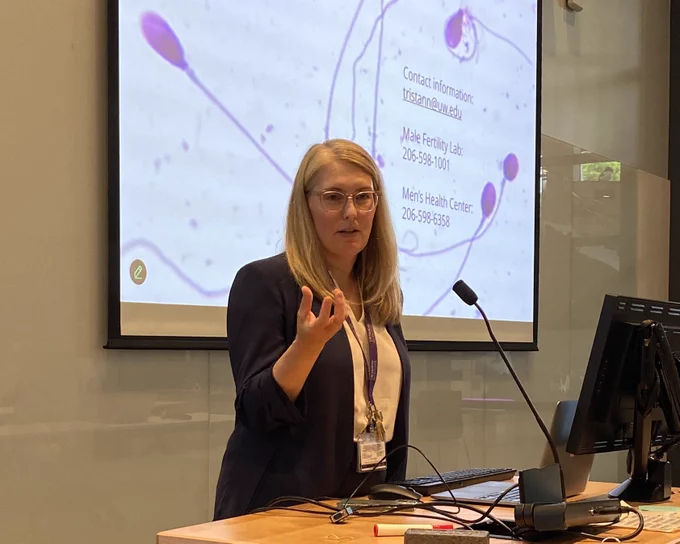Woman with long blond hair and glasses wearing a blue blazer with a projected image of sperm on a screen behind her.