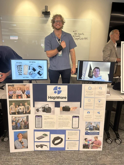 Founder and CEO, Jack Walters, wearing ALeye wristband and glasses behind a HapWare display trifold showing employee pictures and HapWare prototypes. Next to him are two monitors, one monitor showing HapWare prototypes and the other showing a video with different emotions and expressions that the device can decode.