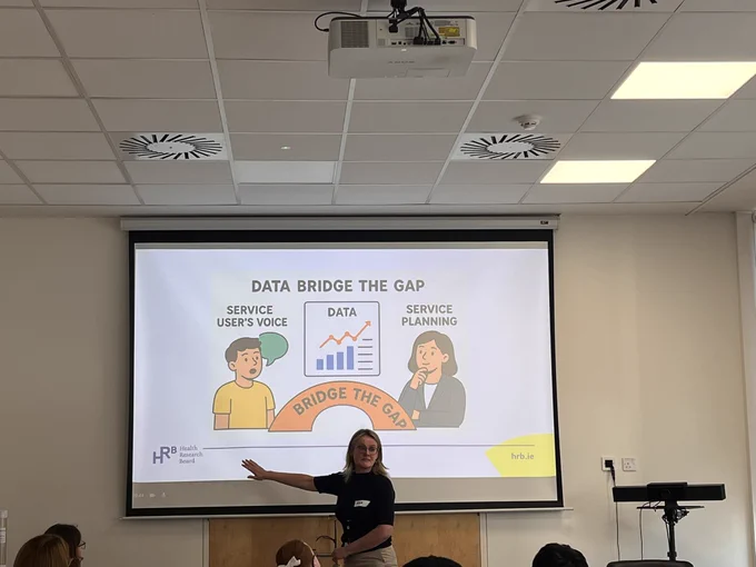 Ena from the HRB pointing to a projector screen. On the screen is a diagram that depicts a young person speaking labelled “service users voice”. There is an orange bridge going from them on the left to an adult female on the right, labelled “service planning”. The bridge between the two is labelled “bridge the gap”. Above the bridge are some symbols to recorder research such as the word data, a line graph, a bar chart and a written document.