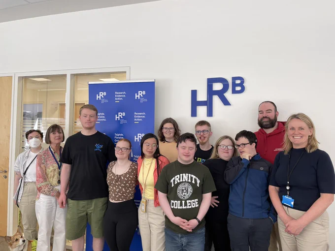1st year TCPID students 4 are male - Alex, Jack, Aidan, Conor, and 4 are female - Makayla, Isabel, Ava, Niamh. Accompanied by two staff from TCPID Angela and Des, and Ena and Mary from HRB. The HRB logo in blue lettering is on the wall behind the group.