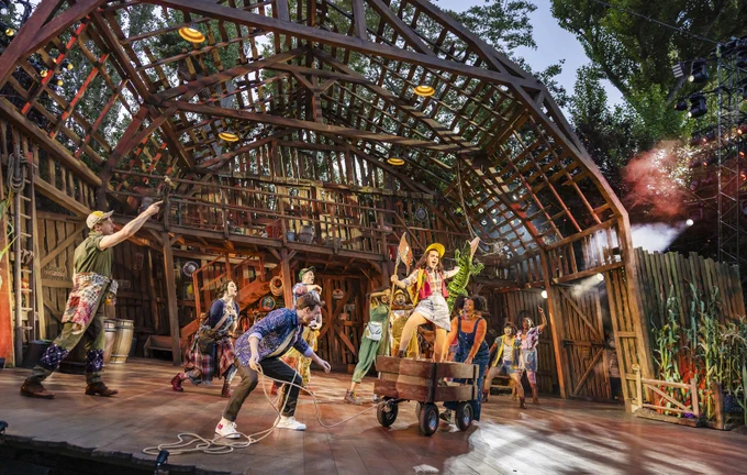 Maizy (performed by Sophie McShera) poses triumphantly on top of a wooden cart, with a Tampa Bay Buccaneers Flag in one hand and a stuffed crocodile in another. The company of Shucked cheer. Behind, a towering barn leans precariously to the left. 