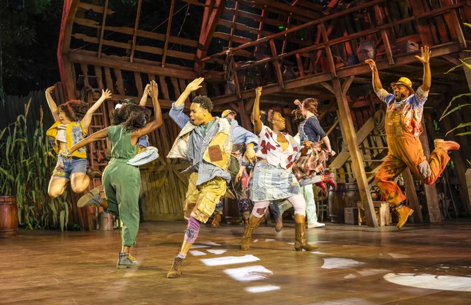 The company of Shucked dance onstage in celebration, a few of them mid-air as they leap. 