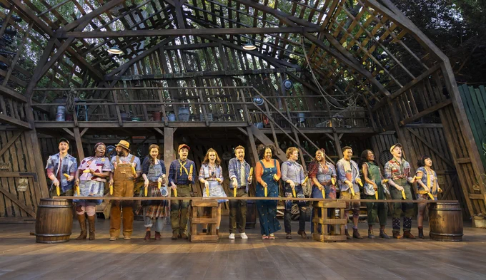 The full company of Shucked stand in a line, singing and dancing in sync with corn in their hands. Behind, a towering barn leans precariously to the left. 