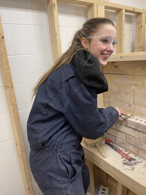 A student smiles and poses for a photo while holding a tool and standing in front of a brick wall being built. 