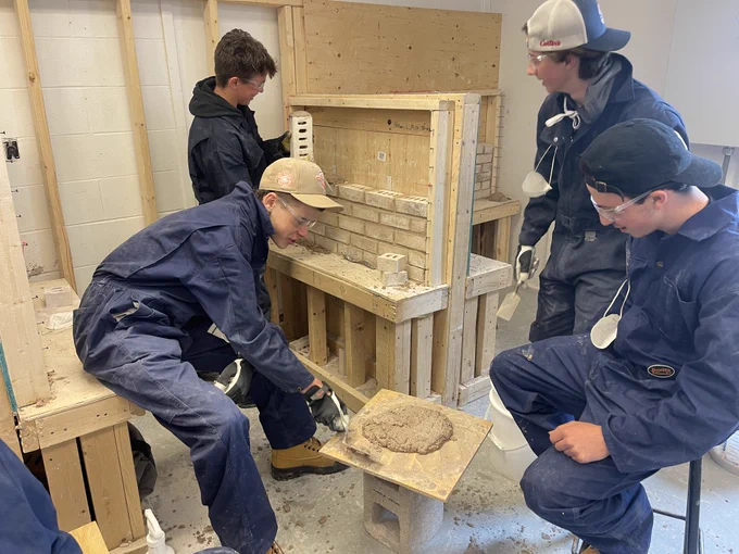 Four students participate in a bricklaying lesson. One student holds a brick while the others hold tools and look on.