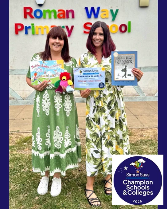 Photograph of Charlotte and Katie. They are stood next to each other, outside on a grass verge in front of an exterior wall. The wall has ‘Roman Way Primary School’ in various primary colour wall mounted letters on it. Charlotte and Katie are both smiling at the camera. They are holding up bereavement story books, a cuddly teddy bear and their Simon Says Champion School certificate. It is a bright day.