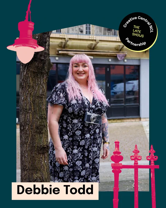 Photograph of commissioned artists Debbie Todd, a photographer with long pink hair, wearing a black floral dress and a vintage camera hangs around her neck. She is standing outside, leaning against a tree with a smile. The background shows a building facade with large windows and a Creative Central NCL and The Late Shows partnership badge in the top right corner.