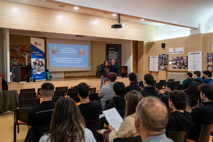 Una sala de conferencias con gente de público sentada y dos personas frente al escenario hablando. 
