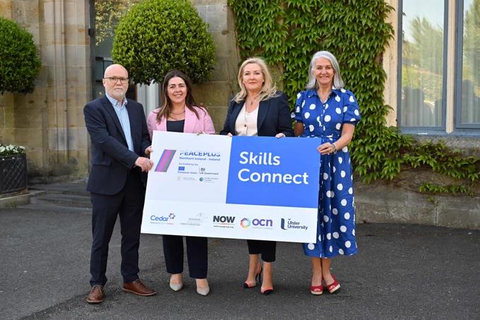 [ID Image shows four PEACEPLUS Skills Connect stakeholders holding large sign. They are smiling at camera in a sunlit courtyard. ]