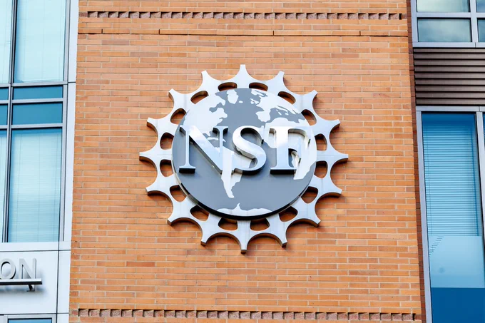 The National Science Foundation logo mounted on a brick wall of the NSF headquarters building, featuring a stylized globe surrounded by gear-like spokes.