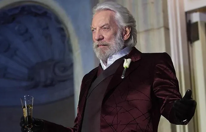 Actor Donald McNichol Sutherland, dressed in a burgundy suit, holding a champagne glass, depicting President Coriolanus Snow in The Hunger Games film series.