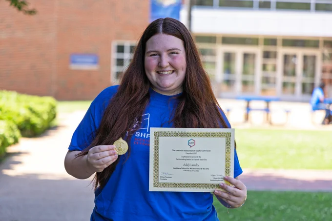 A young woman with long, reddish-brown hair holds a medal and certificate and smiles proudly