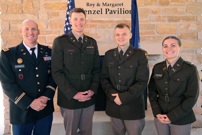 Four military personnel smiling in uniform, standing in front of the Roy & Margaret Kinzel Pavilion sign, with an American flag to the left.