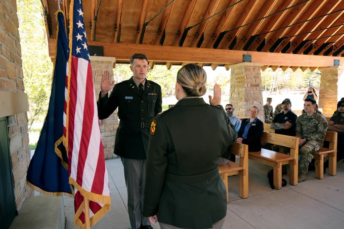 A U.S. military officer is administering an oath under a wooden pavilion, with the American flag displayed to the left. An audience in military and civilian attire is seated in the background.