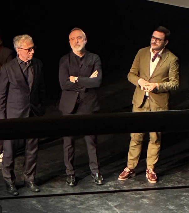 Three individuals are standing on stage, with one wearing a black outfit and the other two dressed in blazers. The middle person has crossed arms and white hair, while the person on the right is adjusting their glasses. The background is dark, focusing attention on the group.