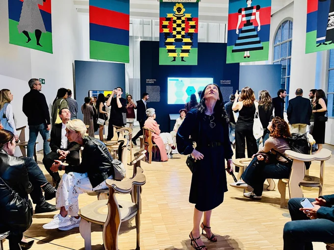 Visitor looking up at an art exhibition at a gallery, surrounded by people and colorful flags hanging on the walls.