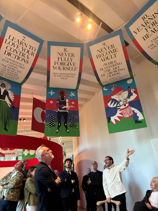 A group of visitors viewing colorful banners hanging from the ceiling at an exhibition, each with text and unique artwork, at the Triennale Milano.