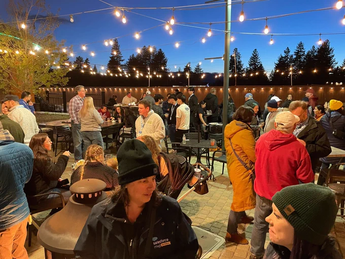 A large group of people mingle outdoors under string lights at a Tahoe Dreamin’ evening social event. Attendees are chatting, wearing warm layers, and gathered around tables and heat lamps.
