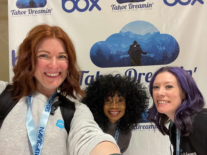 Three smiling attendees pose for a selfie in front of a Tahoe Dreamin’ step-and-repeat banner. They are wearing conference badges and lanyards, and one has a Salesforce logo on her shirt.