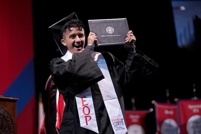 A graduate wearing an EOP stole holds his diploma cover up in celebration as he crosses the stage