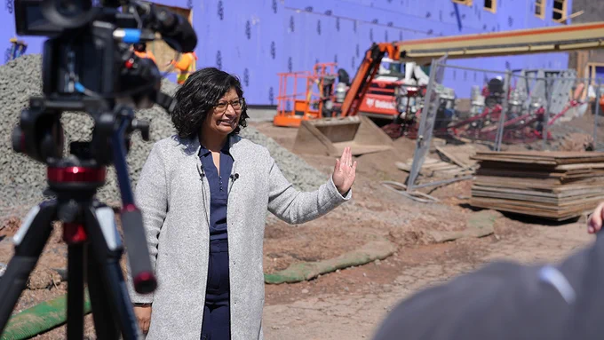 A person wearing a long gray coat waving to someone at a construction site. In the background, heavy machinery and construction materials are visible. A camera is positioned in the foreground, capturing the event.