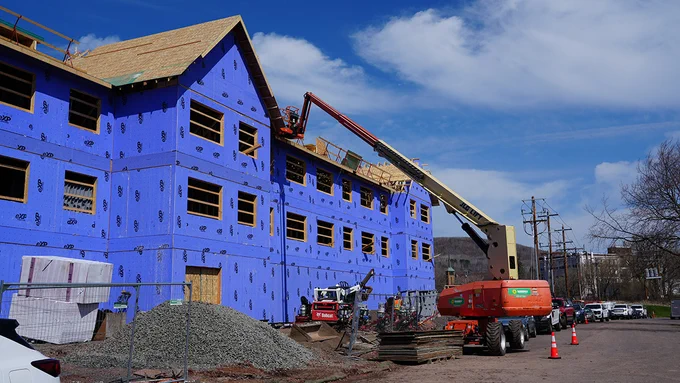 A large building under construction, featuring exposed wooden frames and purple insulation boards. A lift crane is in use and piles of building materials are visible. The scene is set against a clear sky.