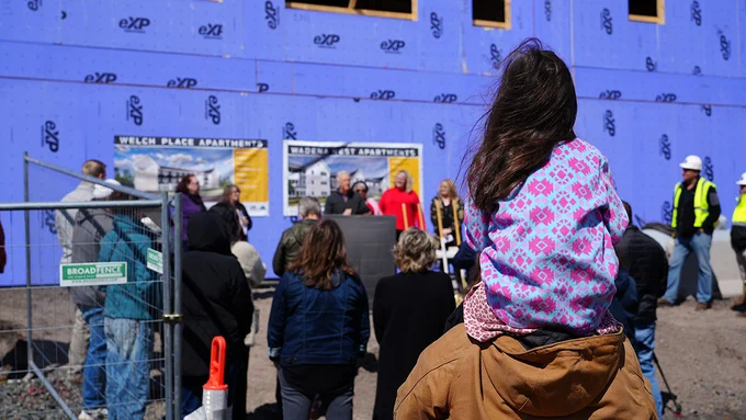A child with long hair sitting atop someone's shoulders viewing a public speech at the construction site of Welch Place Apartments. Several attendees and construction workers are present around a speaker with a microphone.