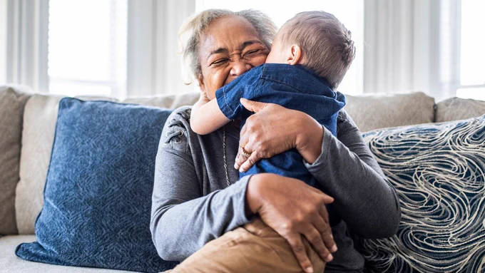A smiling elderly woman embraces a young child on a couch, surrounded by decorative pillows. The setting is bright and comfortable, with natural light coming through the windows.