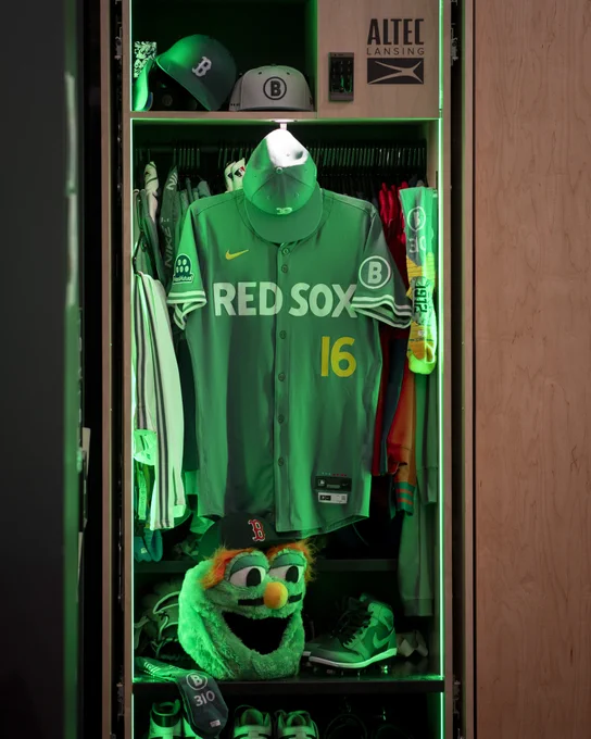 A locked inside the Fenway Park Clubhouse displays the full city connect uniform with the Fenway green jersey hanging with the number 16 and "RED SOX" across the chest. The locker is lit green and the Wally home run head sits at the bottom of the locker. 