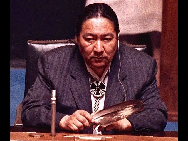 The black-and-white photo shows Elijah Harper seated at a desk, wearing a suit with a patterned tie and a large circular pendant. He holds an eagle feather in one hand, with an earpiece visible, and a microphone on the table in front of him, suggesting a formal or parliamentary setting.
