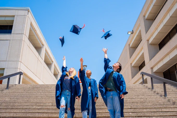 Four graduates throwing their caps in the air on a university staircase, celebrating under a clear blue sky.
