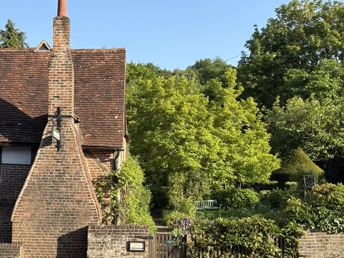 The red-brick chimney and red-tiled roof of Milton's Cottage dominates the left-hand side of this photograph, with the green trees and hedges of the garden to the right, interspersed with vibrant purple iris flowers.